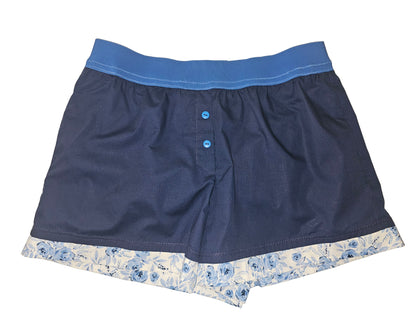 Medium women’s loungewear boxer shorts in deep midnight blue with a 2-inch elastic waistband in a lighter blue shade, finished with soft blue floral trim at the leg opening. Handmade cotton design with deep side pockets for comfortable, stylish lounging.