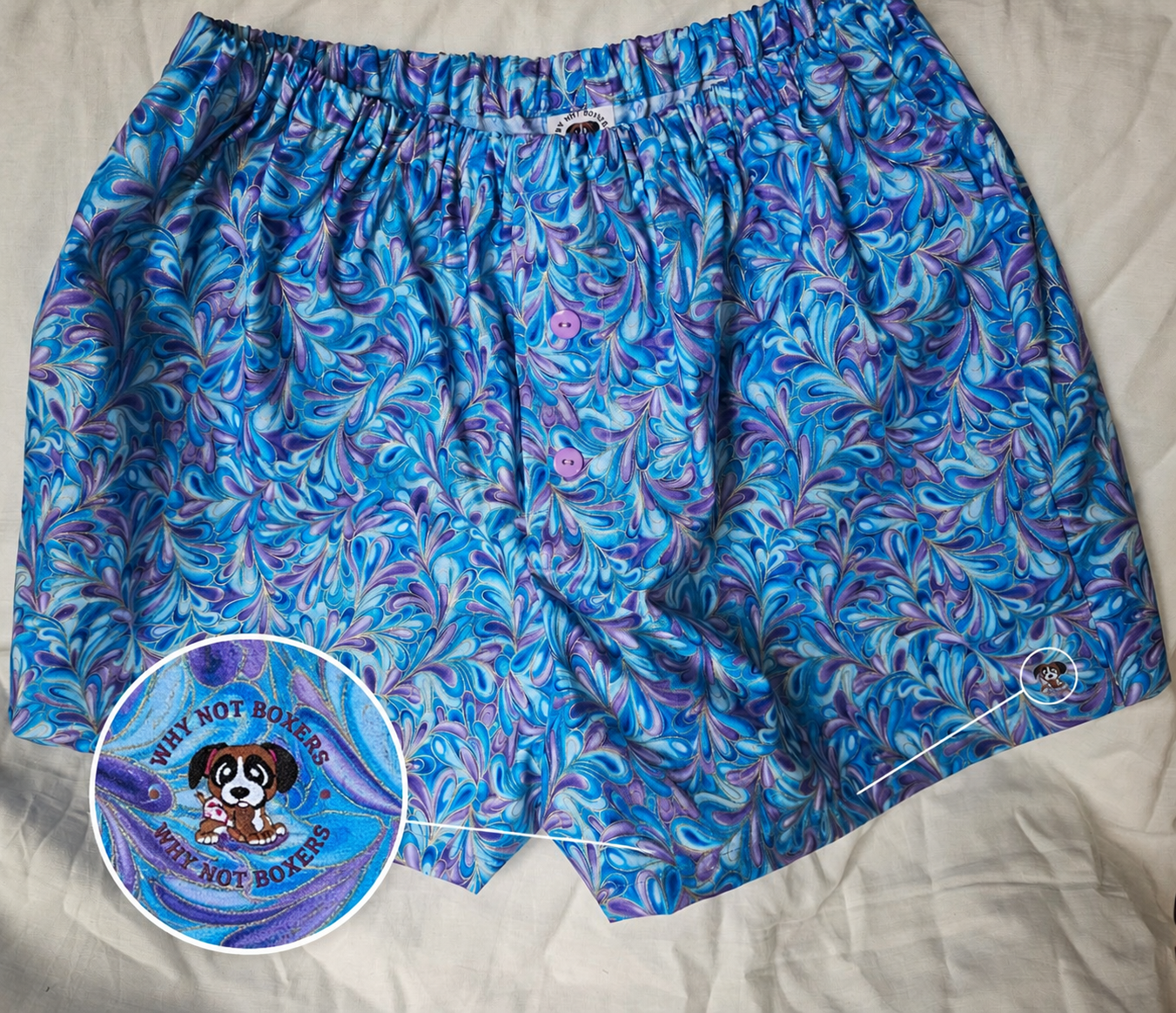 Women’s XL boxer shorts with side pockets, featuring a blue and purple marbled swirl pattern, elastic waistband, and front button detail, displayed on a light background; includes a zoomed inset highlighting the “Why Not Boxers” dog logo printed on the fabric.